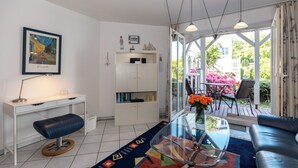 Een moderne woonkamer met een glazen salontafel, een zwarte leren bank, een wit bureau en een boekenkast. Aan de muur hangt een kunstwerk en er is uitzicht op een tuin door grote ramen.