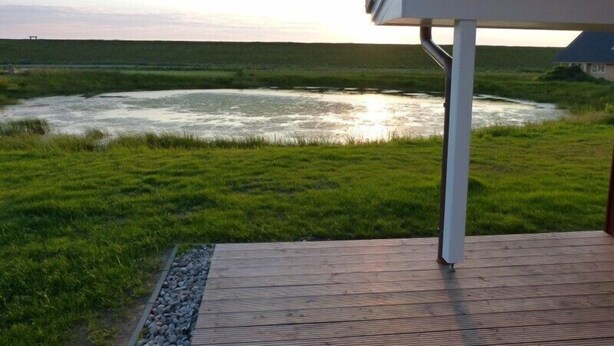 A wooden deck overlooking a river with a grassy bank and a house in the background.
