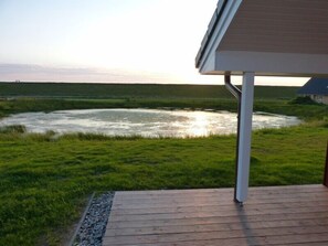 Una terraza de madera con vista a un río, una orilla cubierta de césped y una casa al fondo.