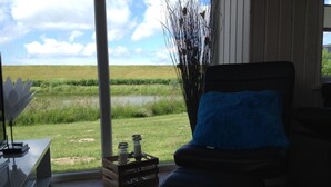 A room with a view of a grassy field and a body of water. There is a black chair with a blue cushion, a wooden crate with candles, and a tall plant in the corner.