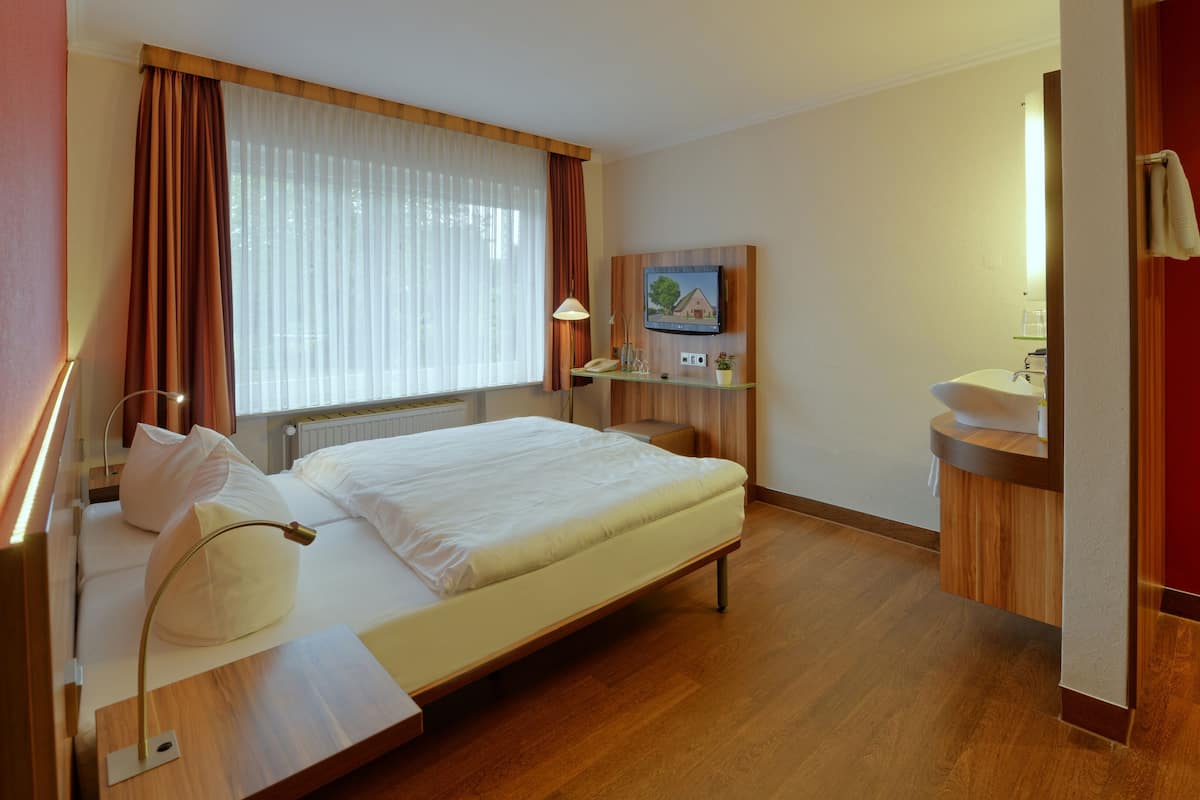 Habitación de hotel con cama, mesita de noche, lámpara, televisor y un baño pequeño con lavabo y espejo.