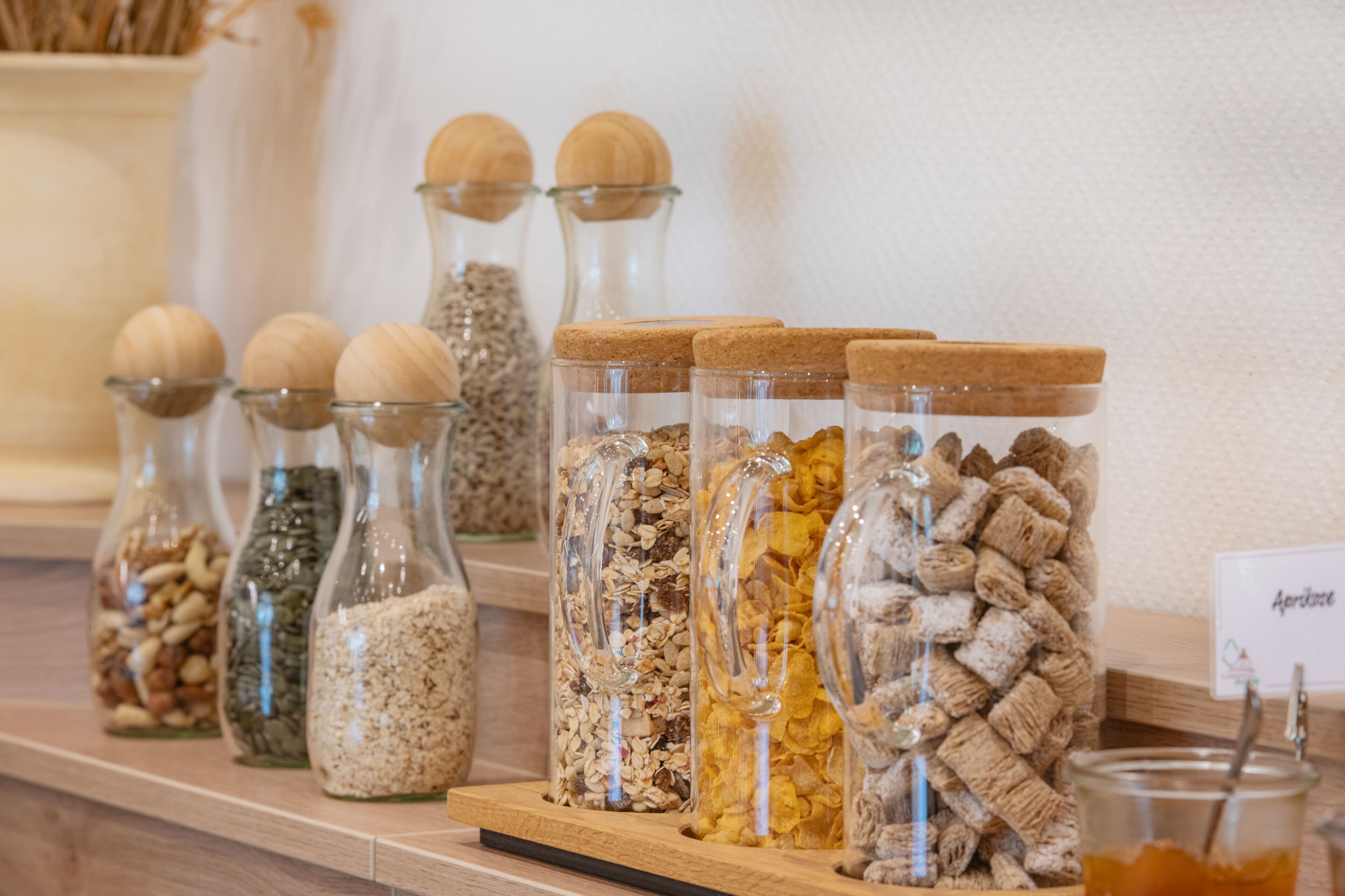 A variety of glass containers with cork lids, filled with different types of grains and seeds, arranged on a wooden shelf.