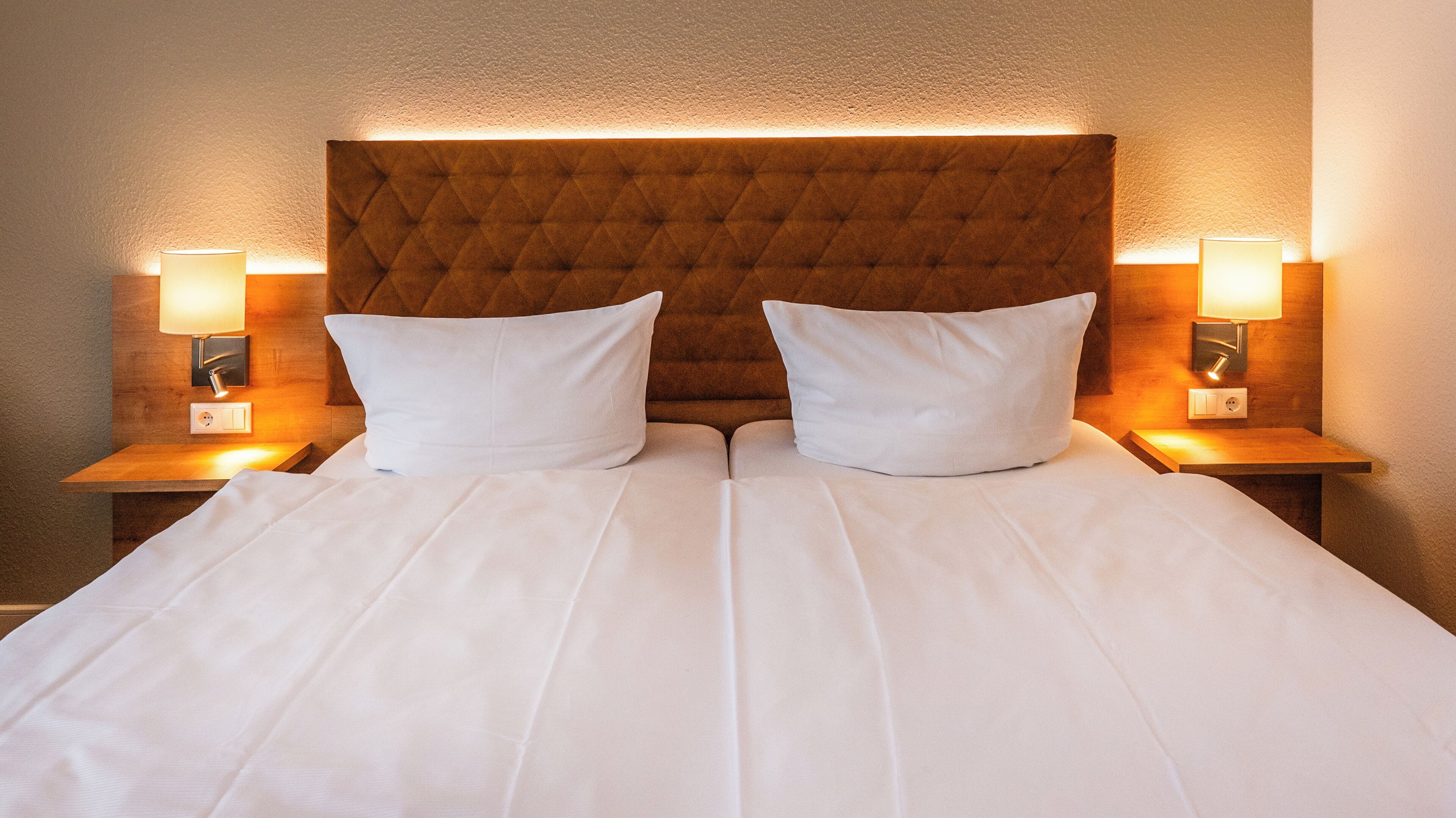Een tweepersoonsbed met een gestoffeerd hoofdeinde, twee bedlampjes en een neutrale wand.