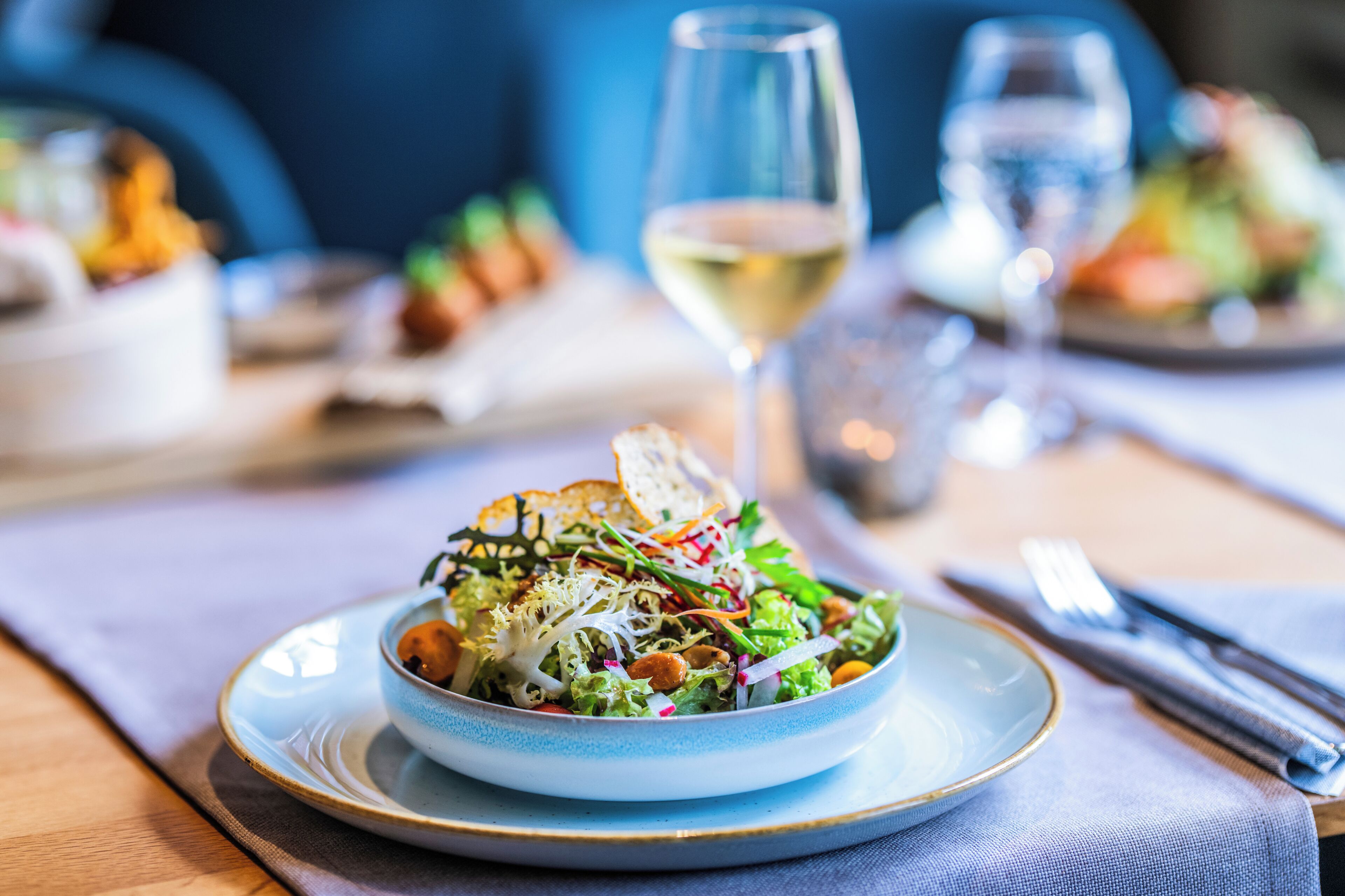 A dining table with a plate of salad, a glass of white wine, and a fork and knife on a napkin.