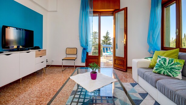 Un salon moderne avec un téléviseur à écran plat, une table basse en verre agrémentée d’une plante, un canapé avec des coussins et une vue sur un espace extérieur, accessible par une porte ouverte.