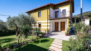 Une maison à deux étages avec un balcon, un jardin où poussent différentes espèces de plantes et un ciel d’un bleu limpide.
