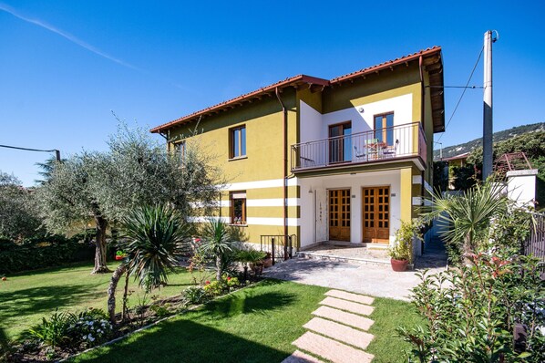A two-story house with a balcony, a garden with various plants, and a clear blue sky.