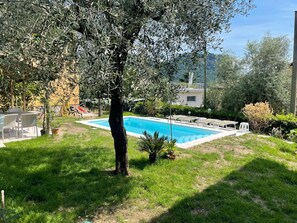 A swimming pool surrounded by grass, a tree, and a building in the background.