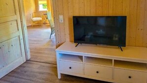 Una habitación con paneles de madera, un televisor sobre un pedestal blanco, una silla y una ventana con luz natural.