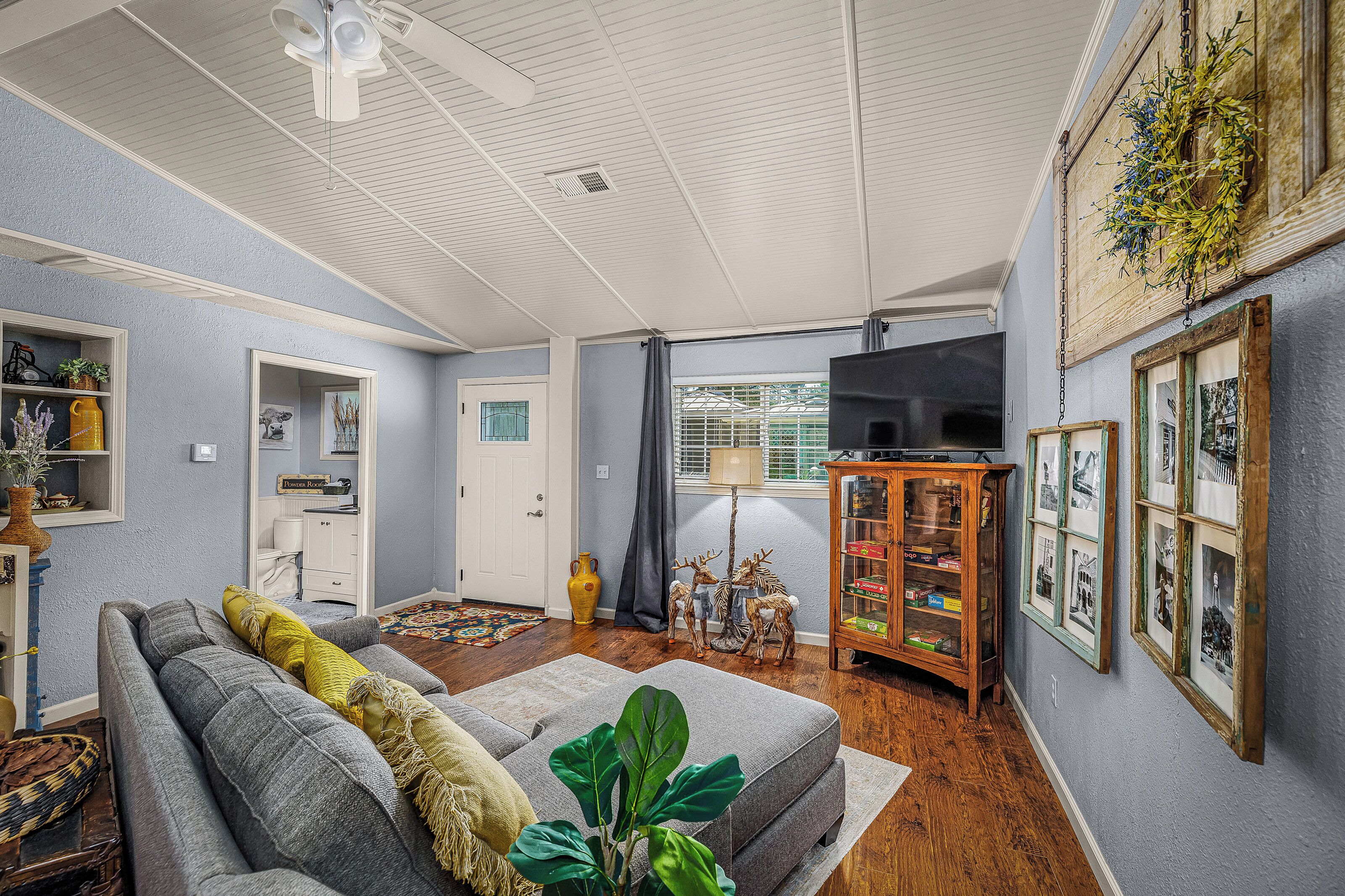 Un salon avec un canapé gris, un meuble en bois orné de cadres photos, une grande plante et un ventilateur de plafond.
