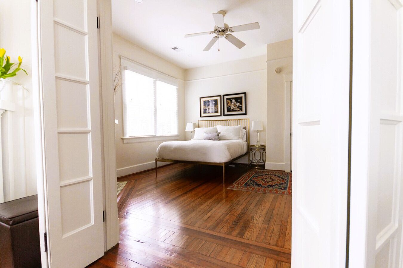 A bedroom with a wooden floor, a bed with white bedding, a ceiling fan, and two framed pictures on the wall.