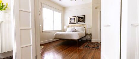 A bedroom with a wooden floor, a bed with white bedding, a ceiling fan, and two framed pictures on the wall.