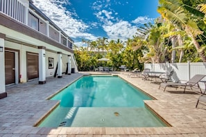 Una piscina rectangular con borde de baldosas, rodeada de sillas de descanso y sombrillas, con palmeras y un edificio al fondo.