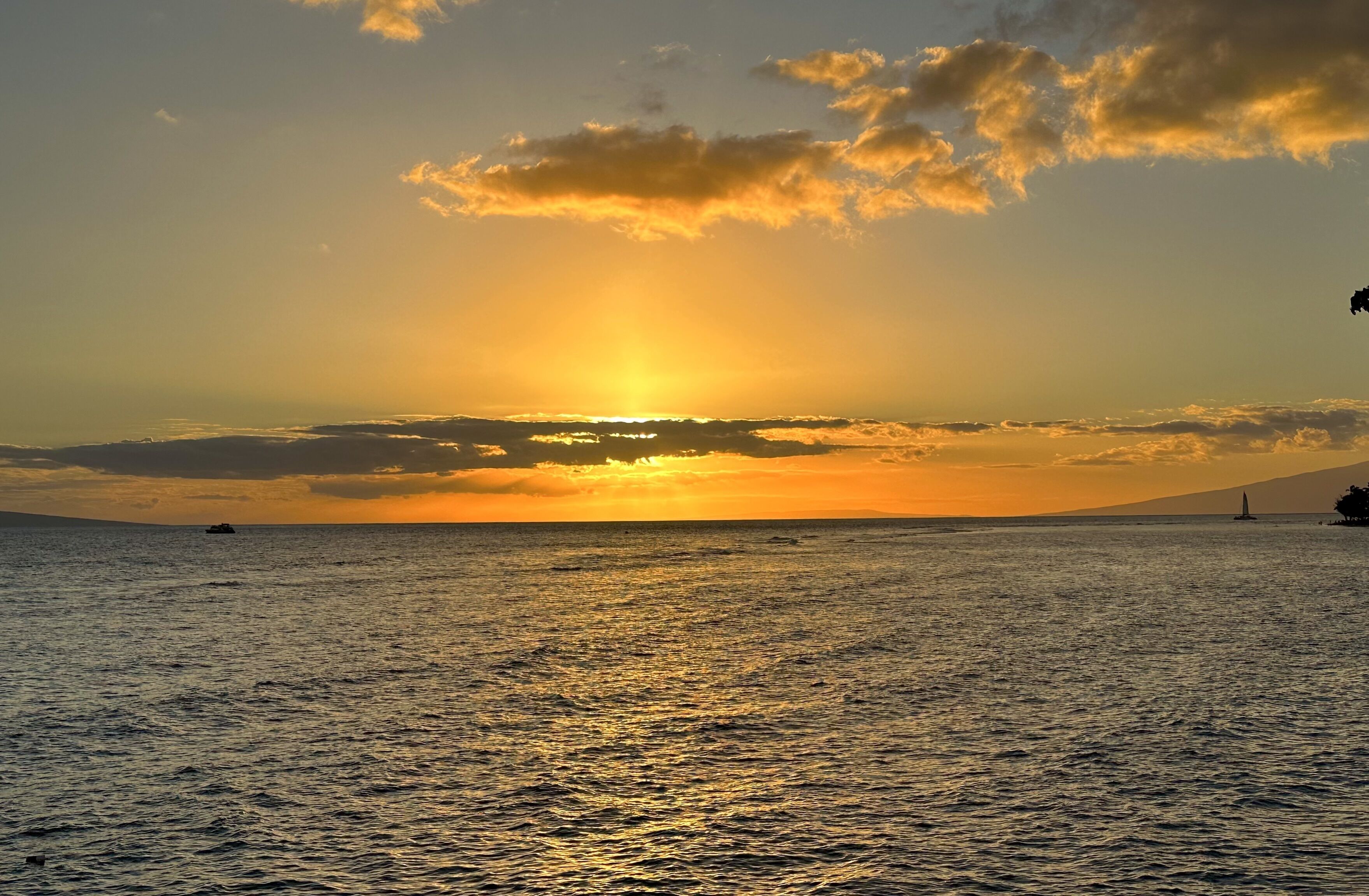 Un coucher de soleil sur une mer calme, avec une terre lointaine à l’horizon et un voilier.
