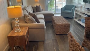 Ein Wohnzimmer mit einer Couch, einem Hocker und einem Fernseher. Es gibt einen Holztisch und eine Lampe.