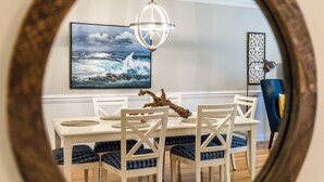 A dining area with a white table and chairs, a painting of a seascape, a chandelier, and a decorative wall piece. - Brass Rail 206 | 30 Yards to Beach, Shared Pool (Tybee Island)
