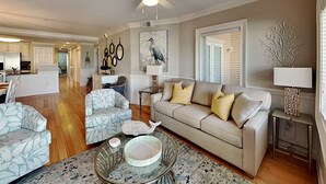A spacious living room with a beige sofa, patterned armchairs, a glass coffee table, and a dining area with a white refrigerator and kitchen counter. - Brass Rail 206 | 30 Yards to Beach, Shared Pool (Tybee Island)