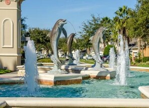 Ein Brunnen mit Delphinskulpturen in einer angelegten Grünanlage mit Palmen und einem Gebäude im Hintergrund.