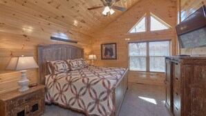 A cozy bedroom with a wooden ceiling, a bed with patterned bedding, a nightstand with a lamp, a dresser, and a ceiling fan.