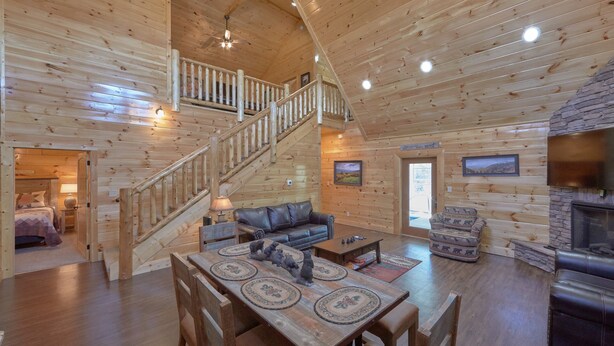 A spacious log cabin interior with a wooden staircase, a fireplace, a dining area, and a living room with a sofa and chairs.