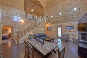 A spacious log cabin interior with a wooden staircase, a fireplace, a dining area, and a living room with a sofa and chairs.