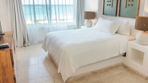 A hotel room with a large bed, two bedside lamps, a view of the ocean, and artwork on the wall.