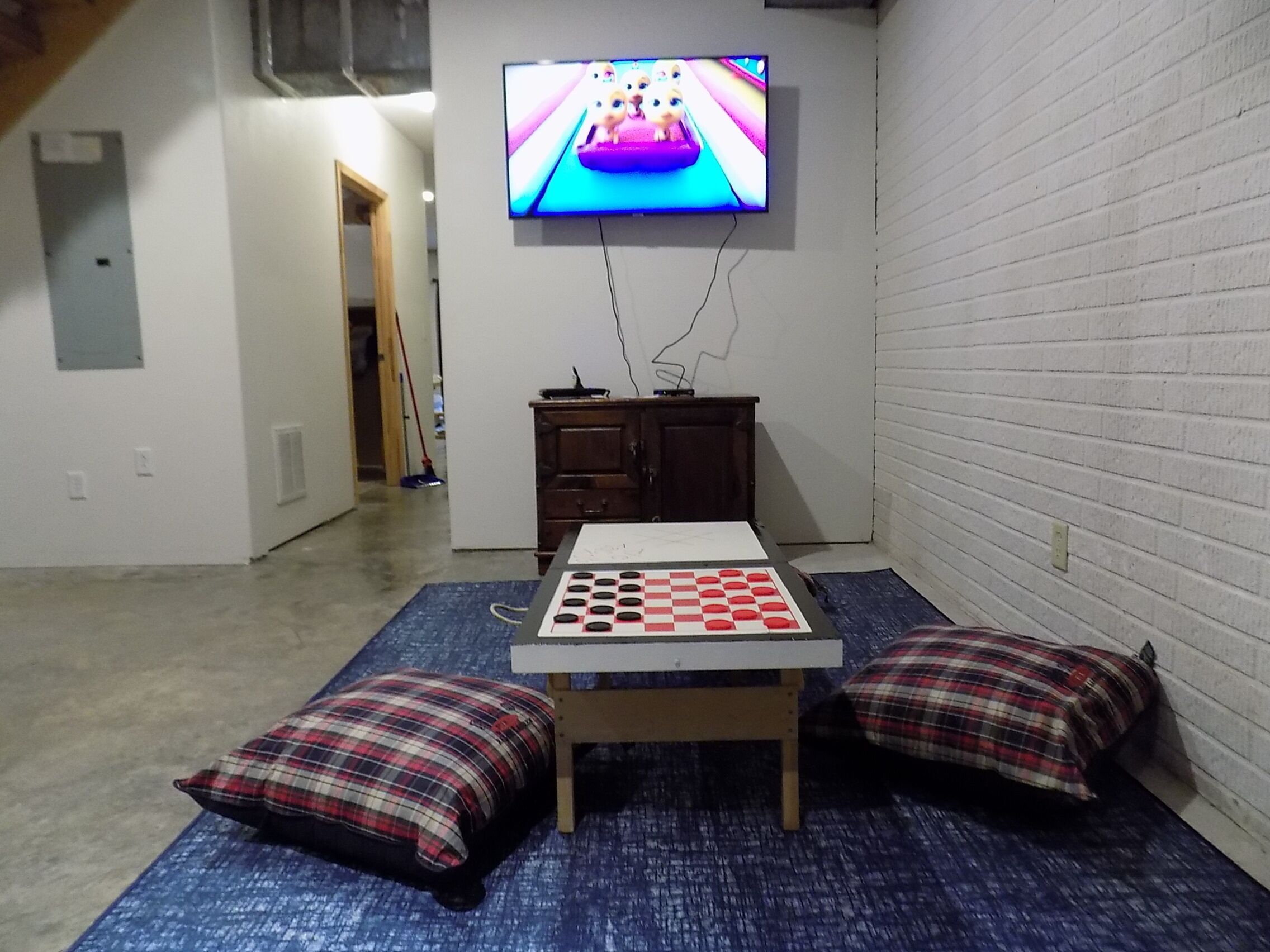 Ett rum med en tv som är monterad på väggen, ett bord med ett schackspel och två kuddar på golvet.