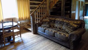 Interior rústico de una cabaña con escalera de madera, sofá estampado, mesa de comedor de madera con sillas y ventana con cortinas.