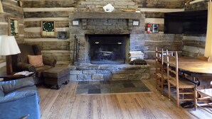 Interior de una cabaña de troncos con chimenea de piedra, un televisor montado en la pared y un comedor con mobiliario de madera.