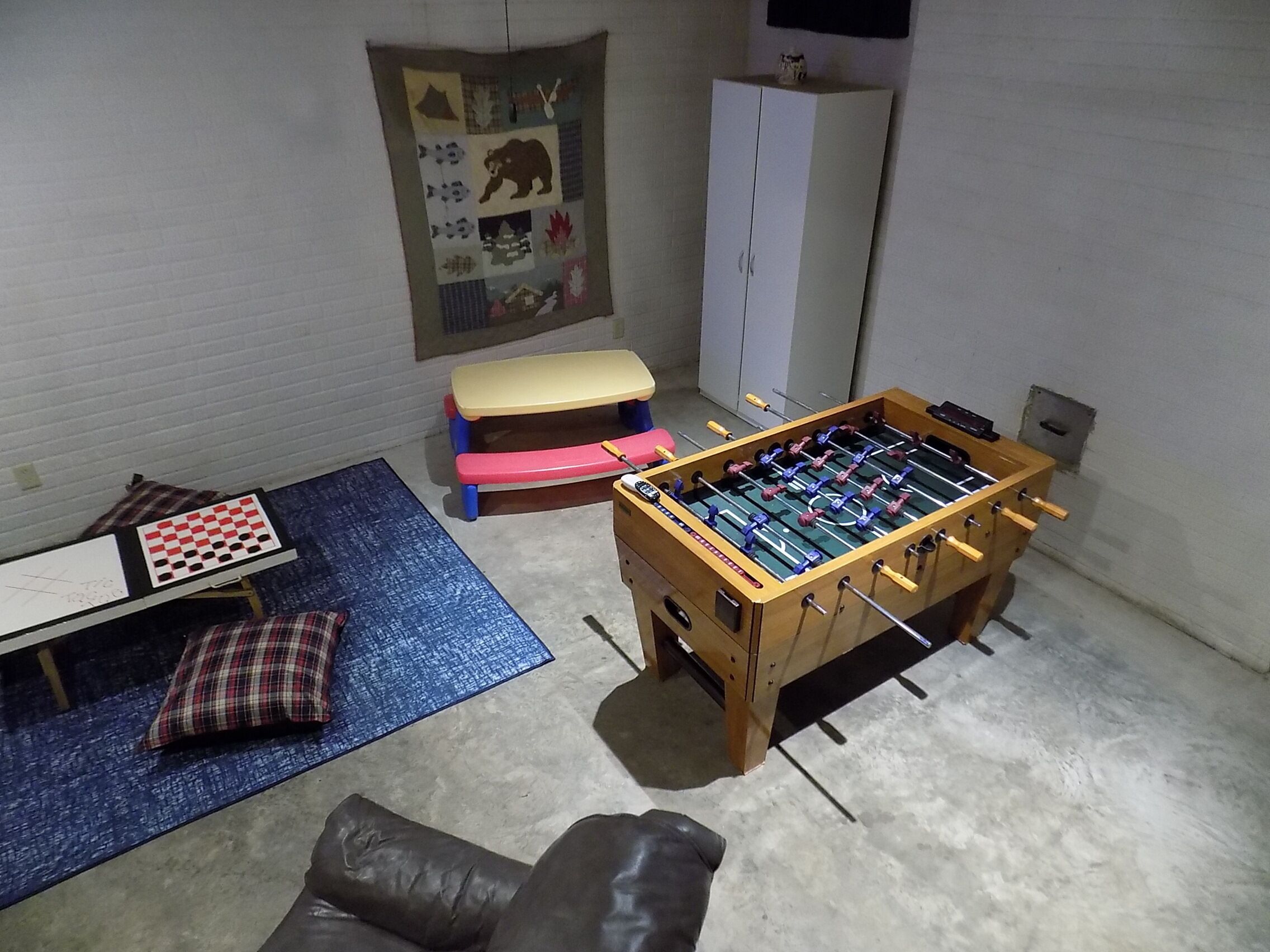 Een kamer met een tafelvoetbaltafel, een bank, een klein tafeltje met geruit tafelkleed en een prikbord aan de muur.