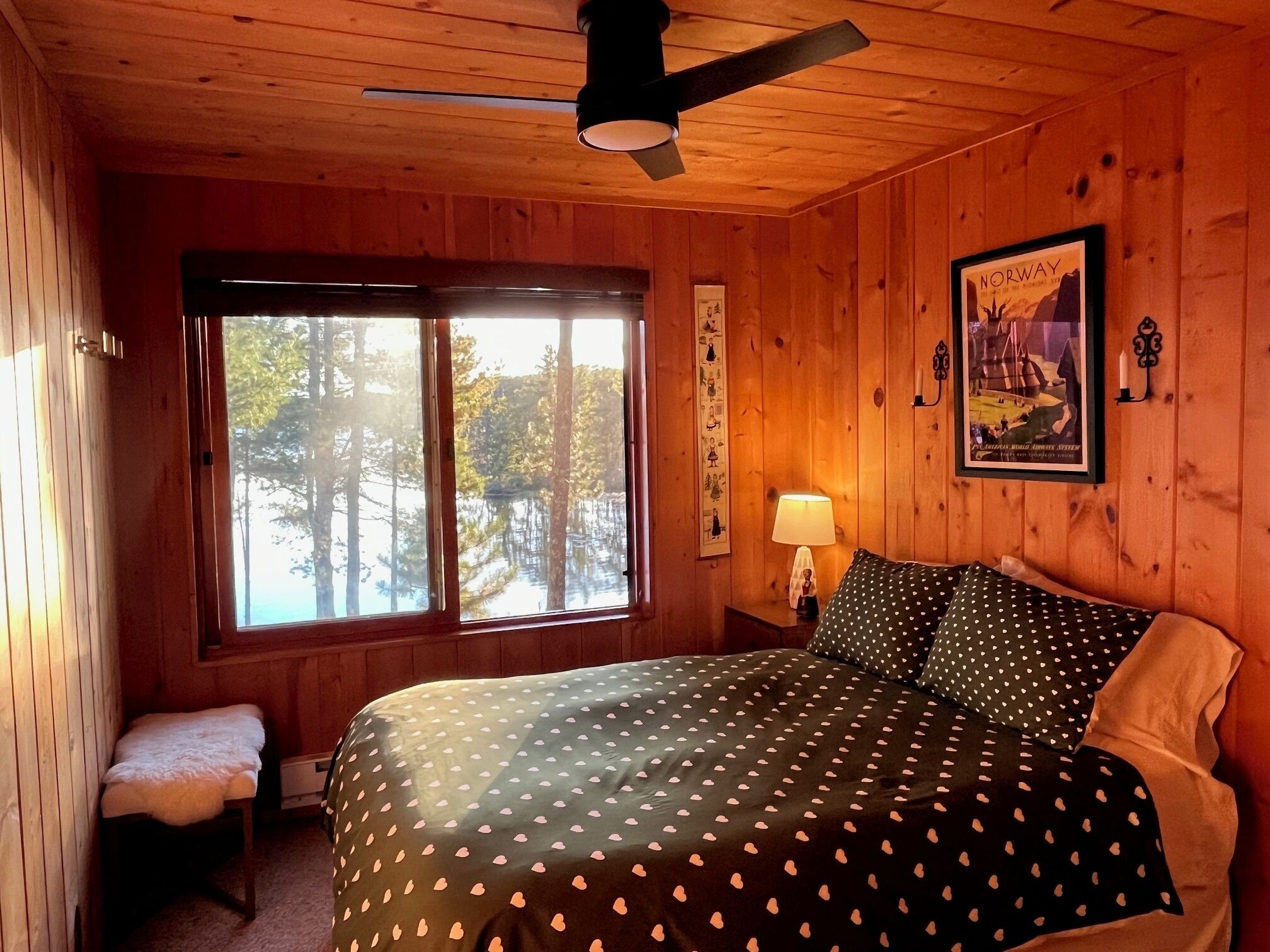 A wooden cabin room with a bed, a bench, a window with a view of trees, a lamp, and a framed picture on the wall.