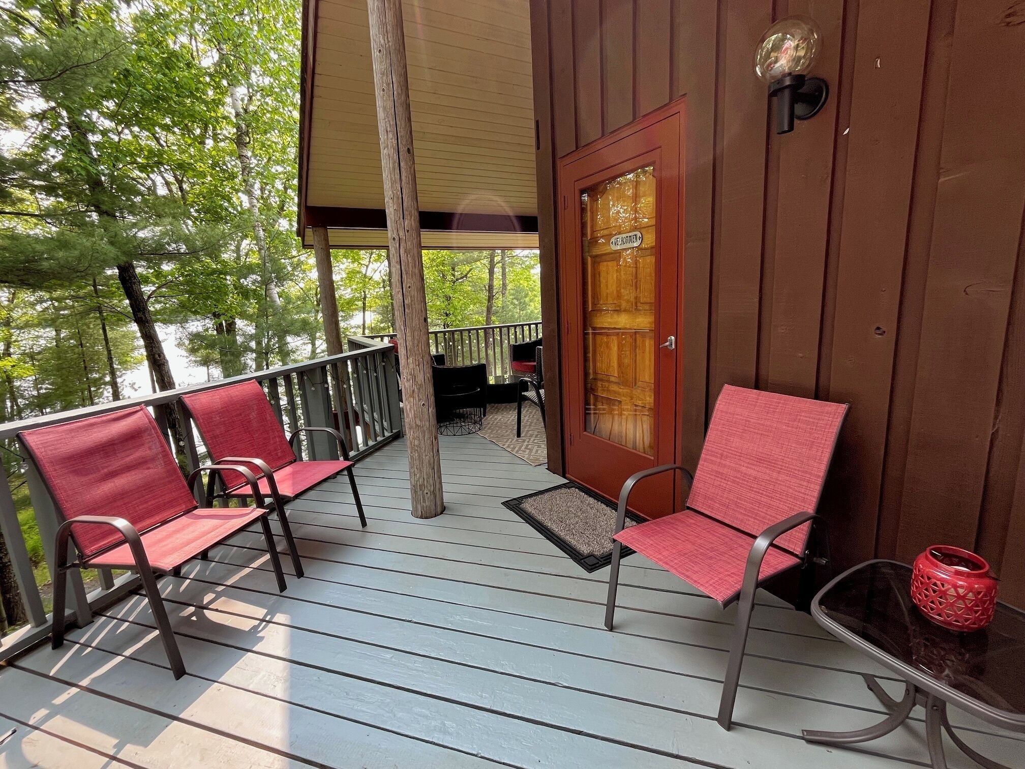 A wooden deck with two red chairs and a small table, a glass door, and a view of trees.