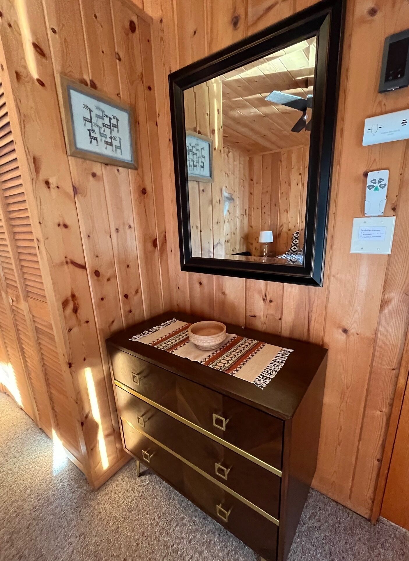 A wooden cabin interior with a wooden dresser, a mirror, and a framed picture on the wall.