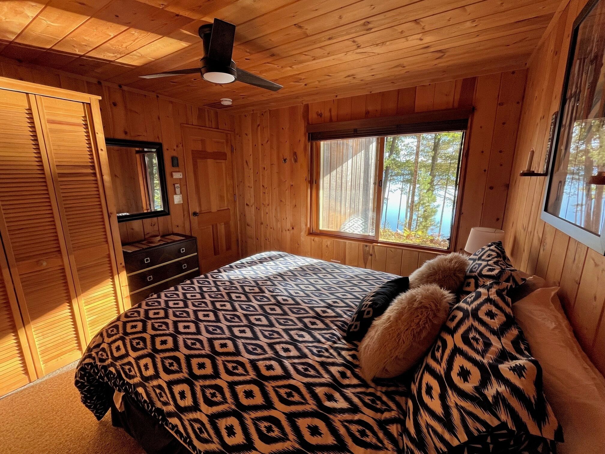 Une chambre dans une cabane en bois, avec un lit, une commode, un ventilateur de plafond et une fenêtre donnant sur la forêt.