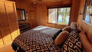 Un dormitorio de cabaña de madera con una cama, un aparador, un ventilador de techo y una ventana con vistas a árboles.