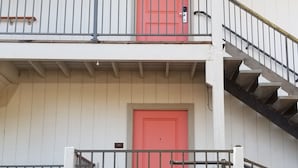 Un immeuble de deux étages avec une porte rouge, un balcon doté d’une rambarde en métal et un escalier menant à l’étage supérieur.