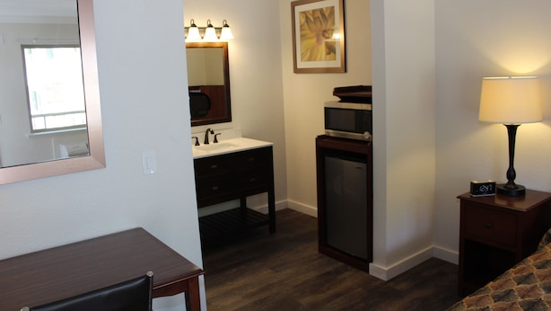 Une chambre d’hôtel équipée d’un bureau, d’une petite kitchenette, d’un miroir avec éclairage, d’une table de chevet avec une lampe et d’une horloge.