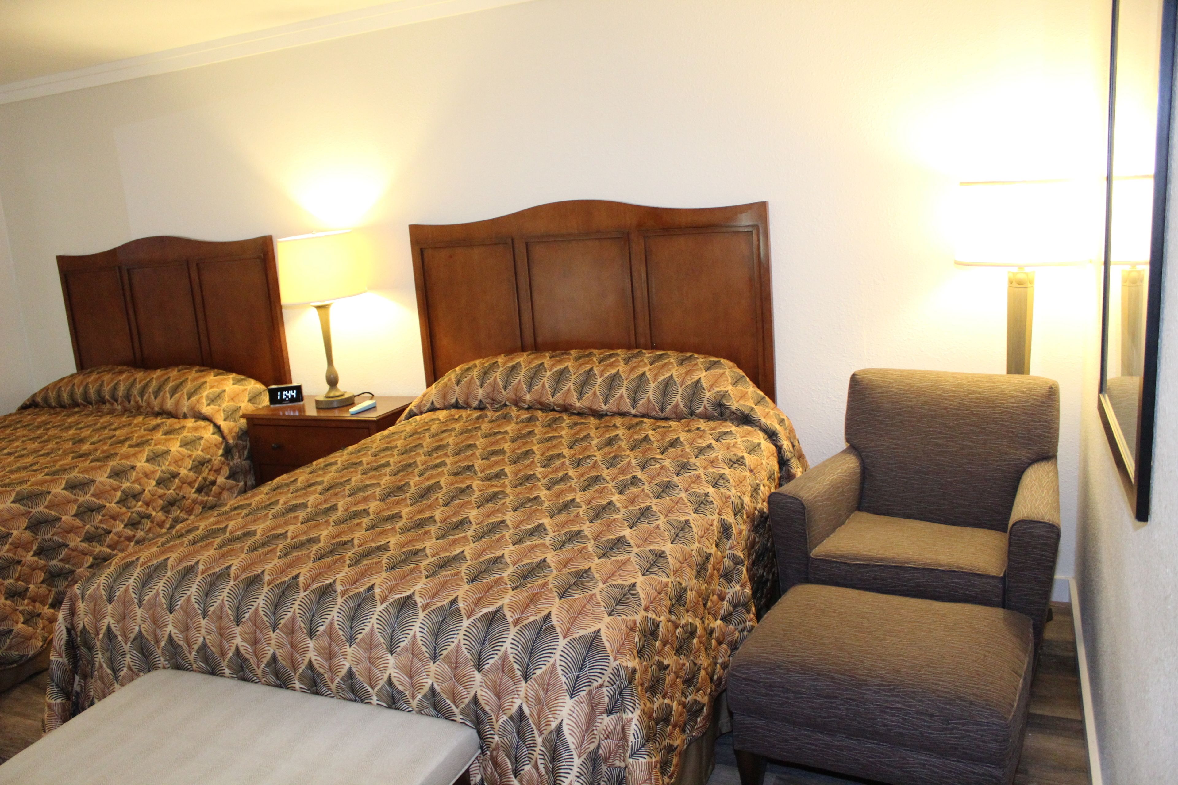 A hotel room with two beds, a nightstand, a chair, and a bedside table.