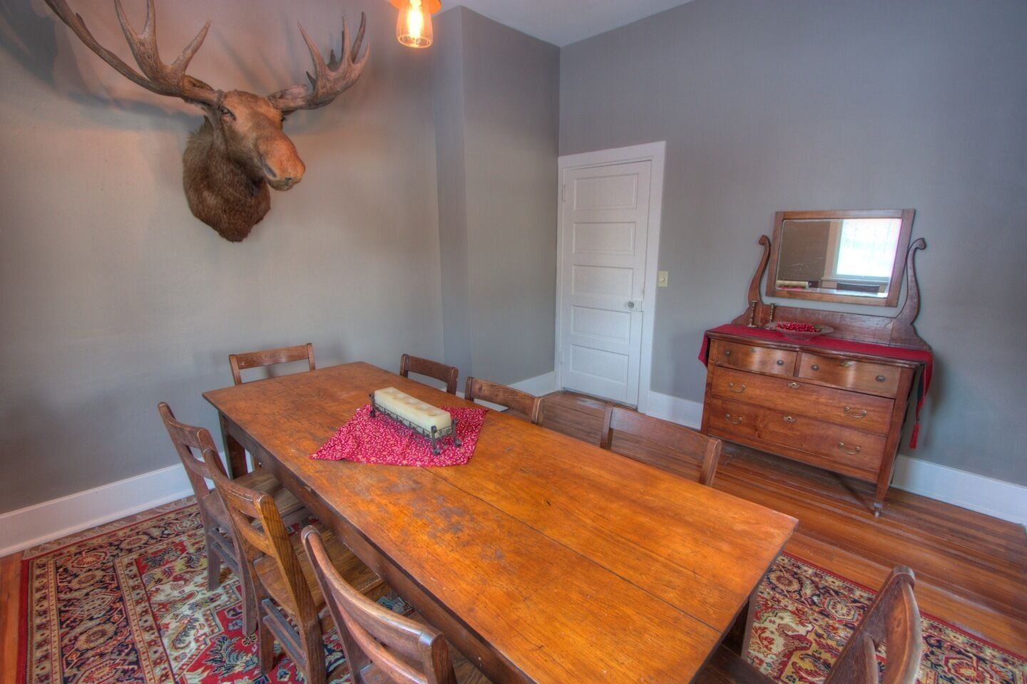 Une salle à manger avec une table en bois, des chaises, un set de table à motifs floraux rouges, une commode en bois et une tête de cerf fixée au mur.