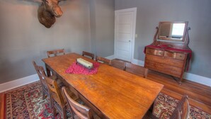 Een eetkamer met een houten tafel, stoelen, een rode placemat met bloemen, een houten dressoir en een aan de muur bevestigd hertenhoofd.