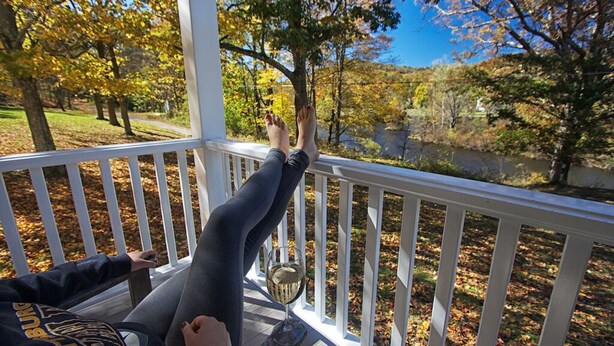 Iemand ontspant op een houten terras met een glas wijn, met uitzicht op een rivier en herfstkleuren.