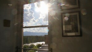 Una habitación con un ventanal amplio que da a un río y montañas, enmarcado por una barandilla y un cuadro colgado en la pared.