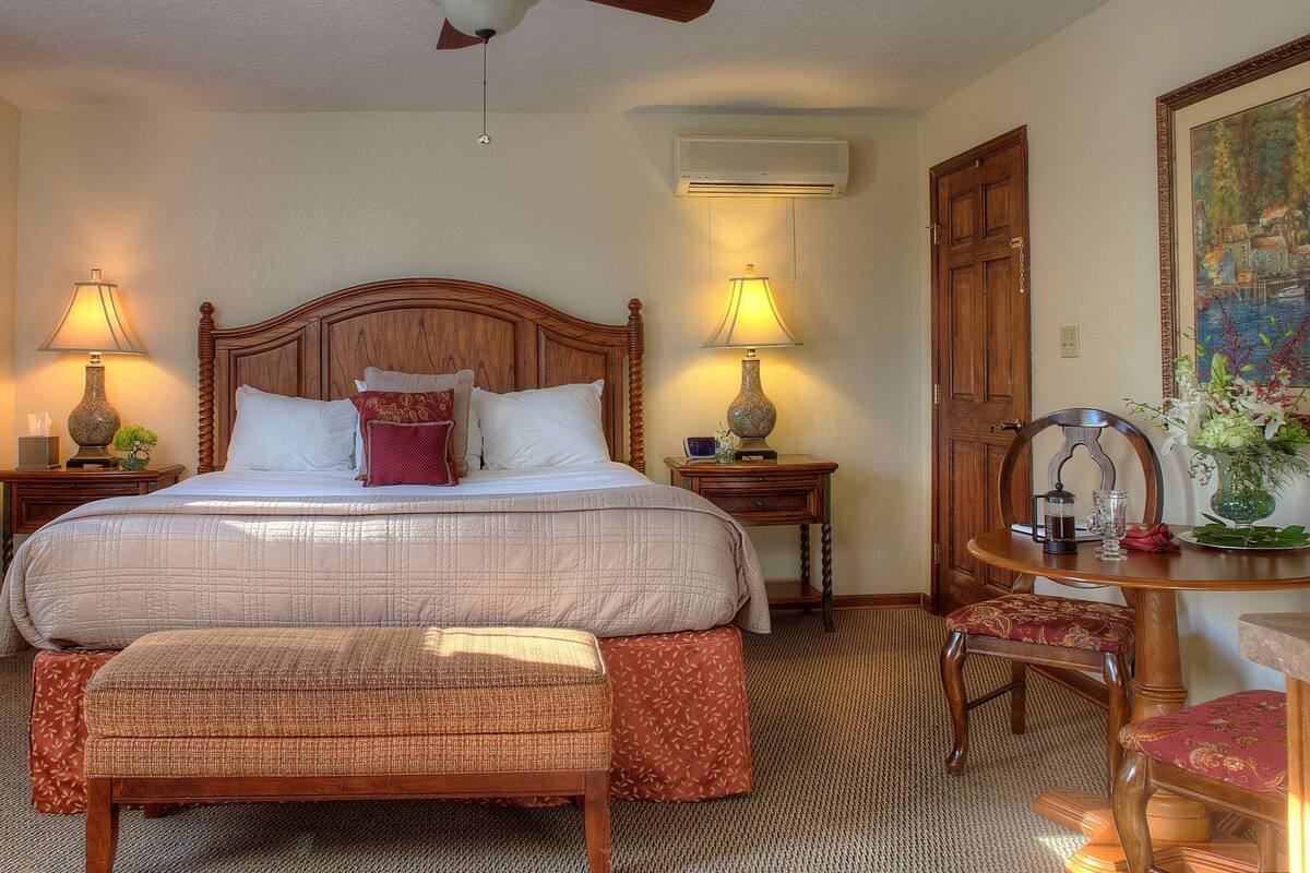 Habitación de hotel con una cama grande, dos lámparas de noche, cabecero de madera, una mesita con un florero y una silla.
