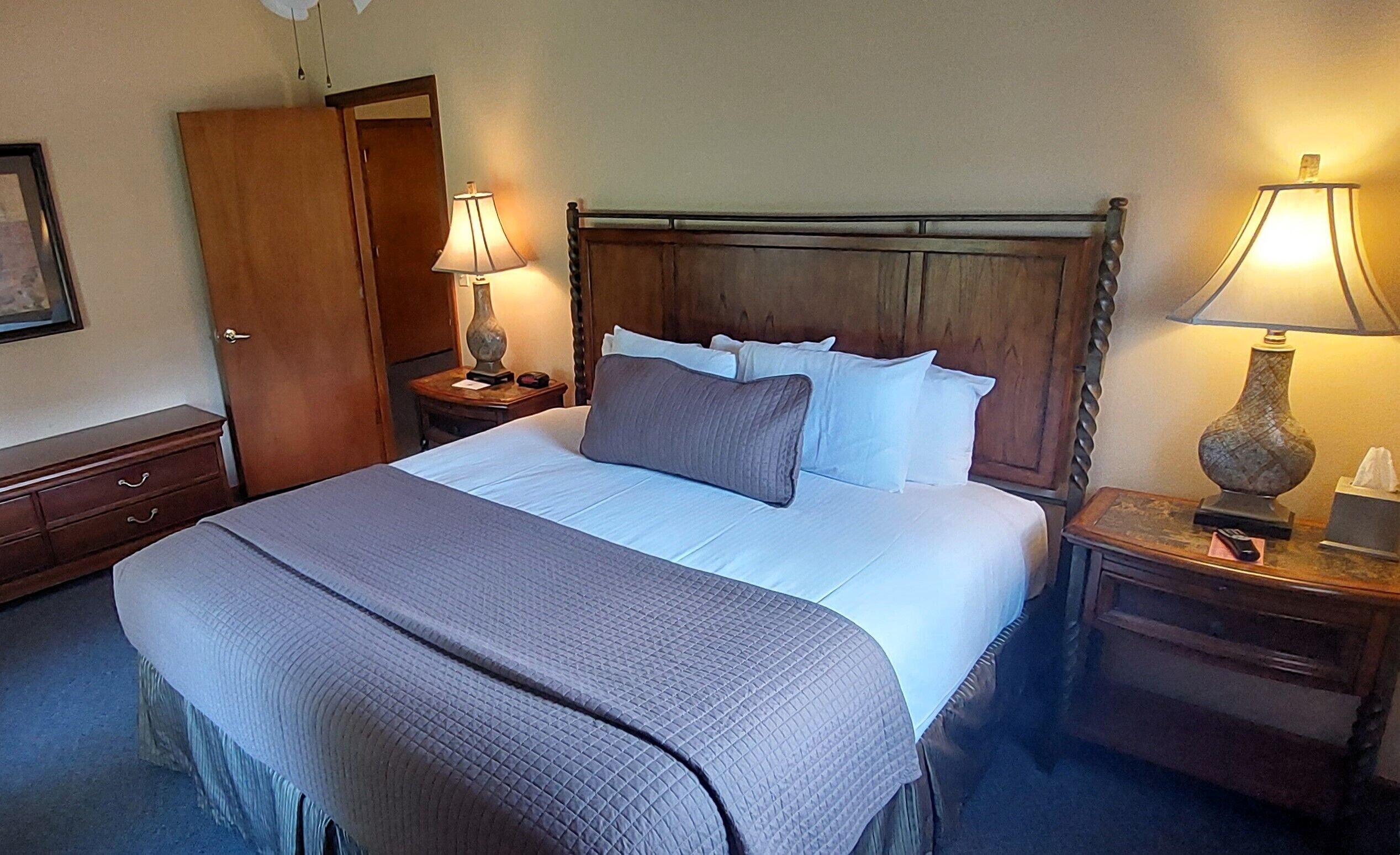 Una cama bien hecha con cabecera de madera, dos lámparas de noche, una mesita de noche de madera y un armario de madera.