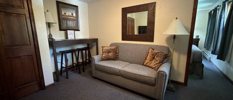 A living room with a sofa, two barstools, a desk, a mirror, and a door.