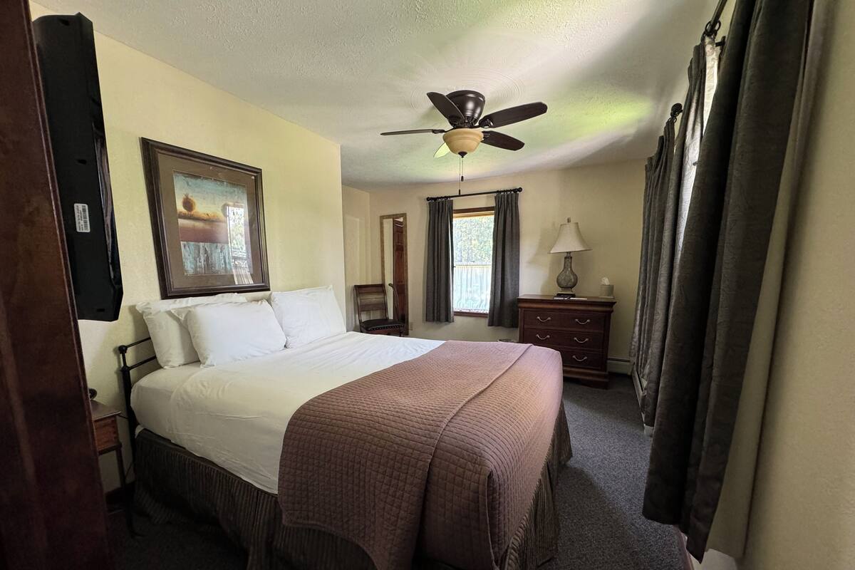 Habitación de hotel con una cama grande, ventilador de techo, un cuadro, una ventana con cortinas, una mesita de noche y una lámpara.
