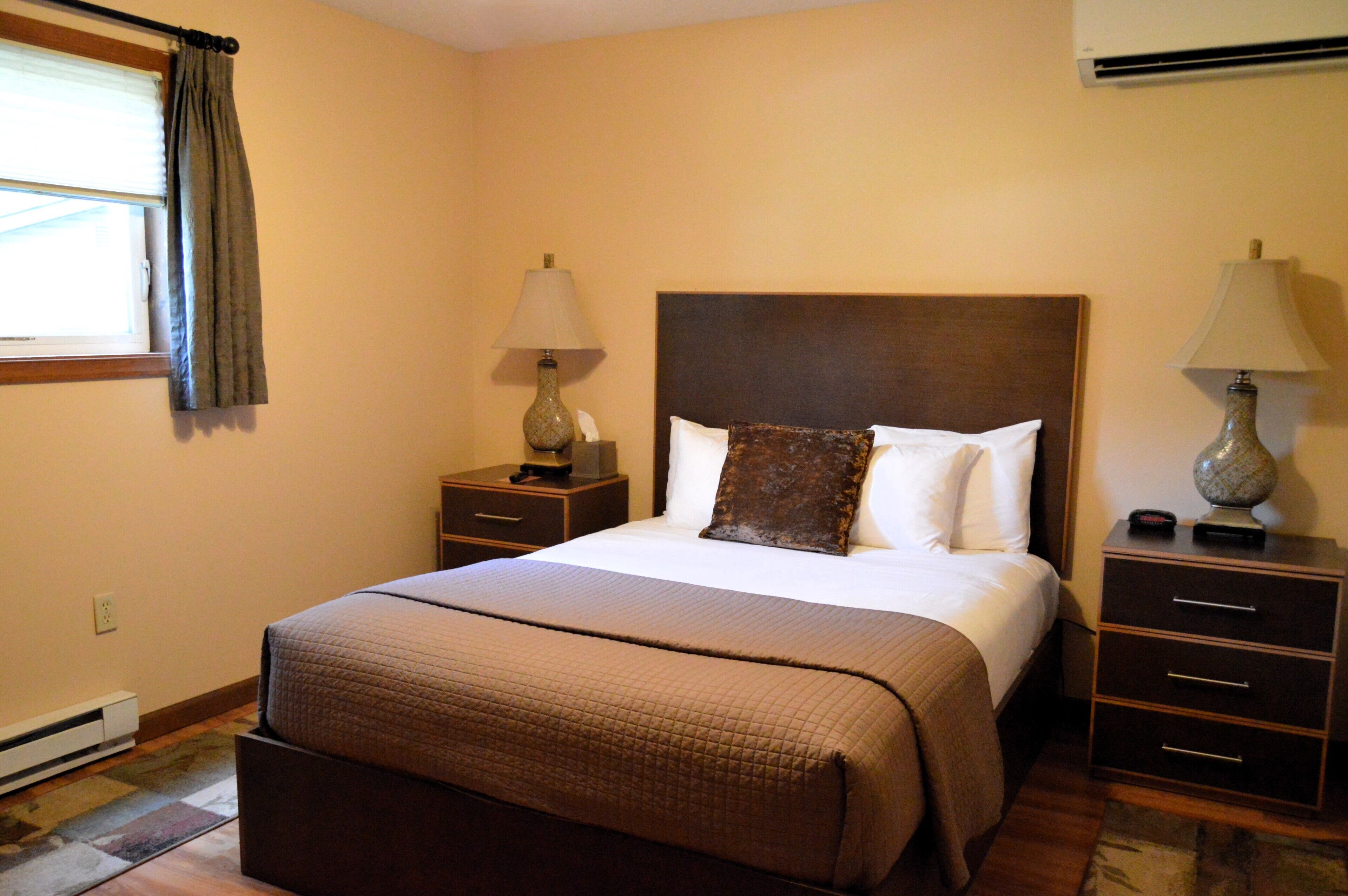 Habitación de hotel con una cama grande, una mesita de noche con lámpara, un aparador con cajones y una ventana con cortinas.