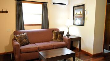 Un salón con un sofá marrón, dos almohadas, una mesa de centro de madera, una lámpara y un cuadro en la pared.