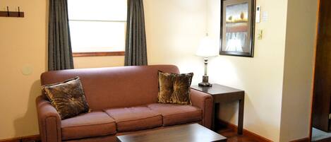 A living room with a brown sofa, two pillows, a wooden coffee table, a lamp, and a framed picture on the wall.