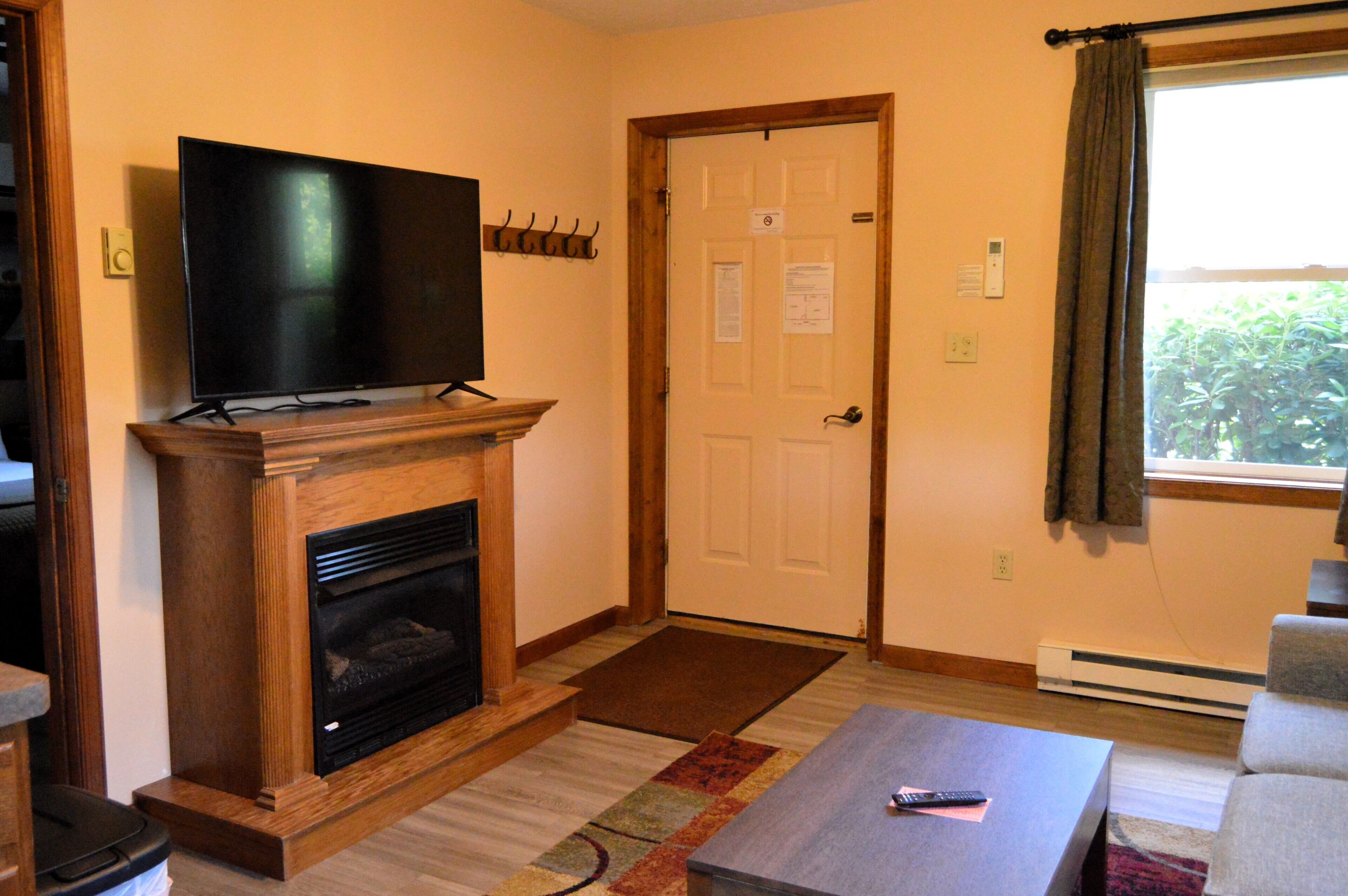 A living room with a fireplace, a flat-screen TV mounted above it, a door with a doormat, and a window with curtains.